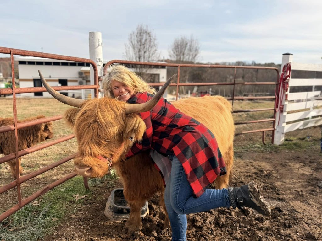 A woman in a red plaid shirt hugs a brown Highland cow in a fenced pasture.